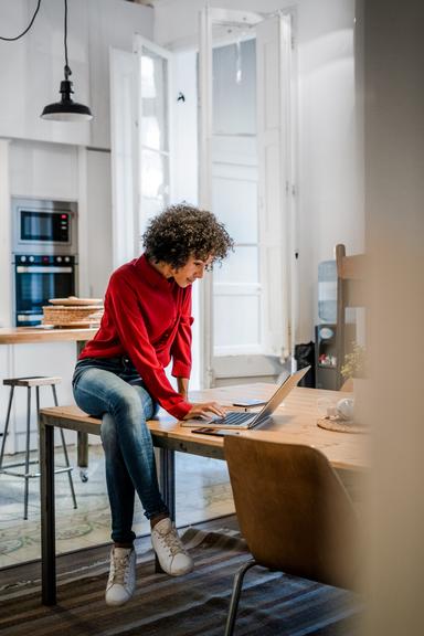 Vrouw met rode trui zit op eettafel en bekijkt iets op haar laptop