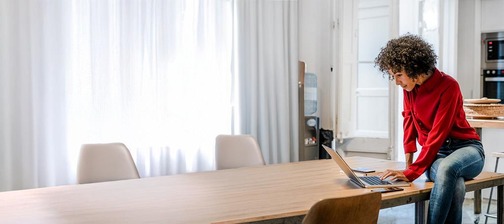 Vrouw met rode trui zit op eettafel en bekijkt iets op haar laptop