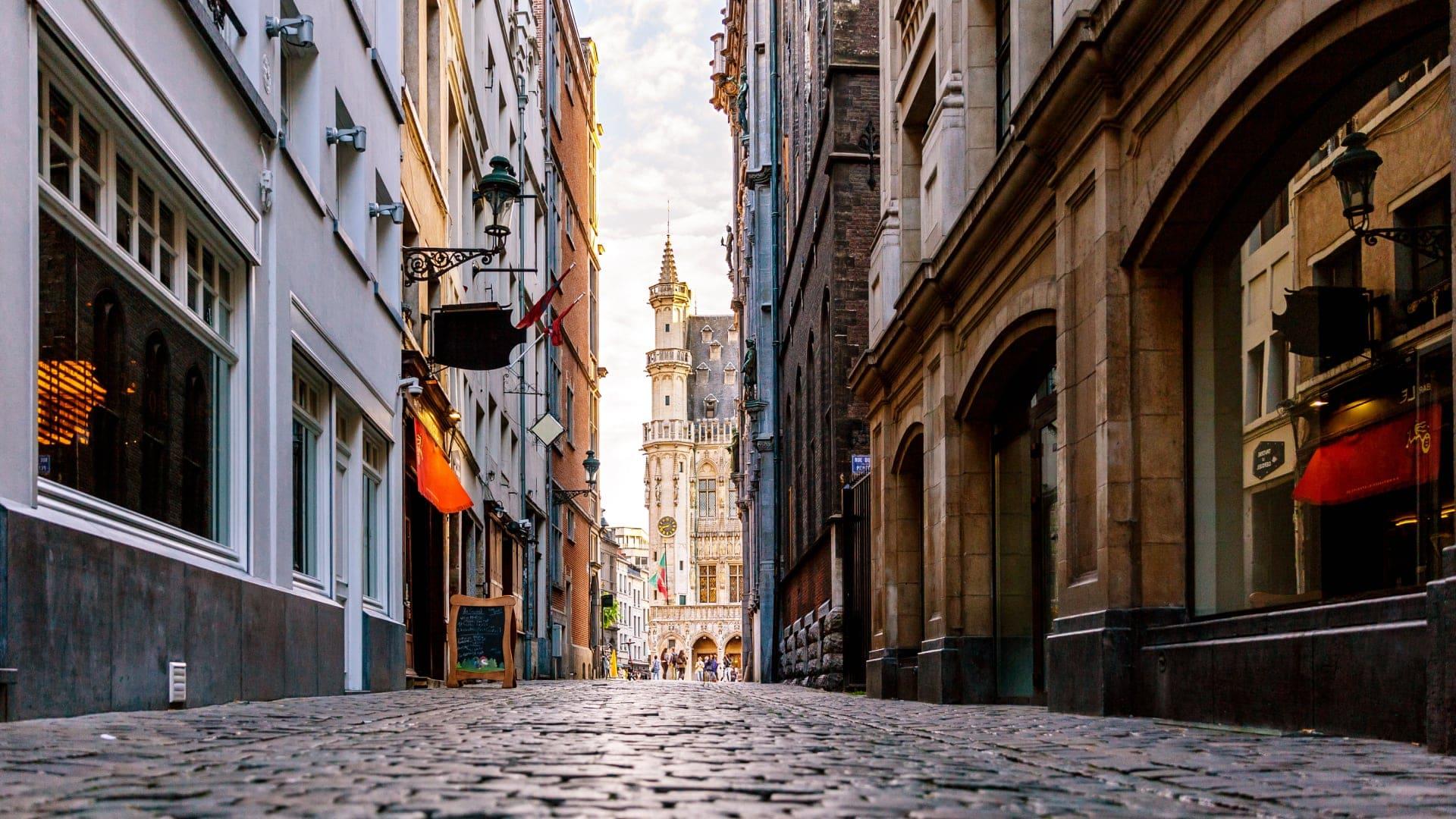 Cobblestone street leading to a grand historic tower amidst Belgian buildings at dusk.